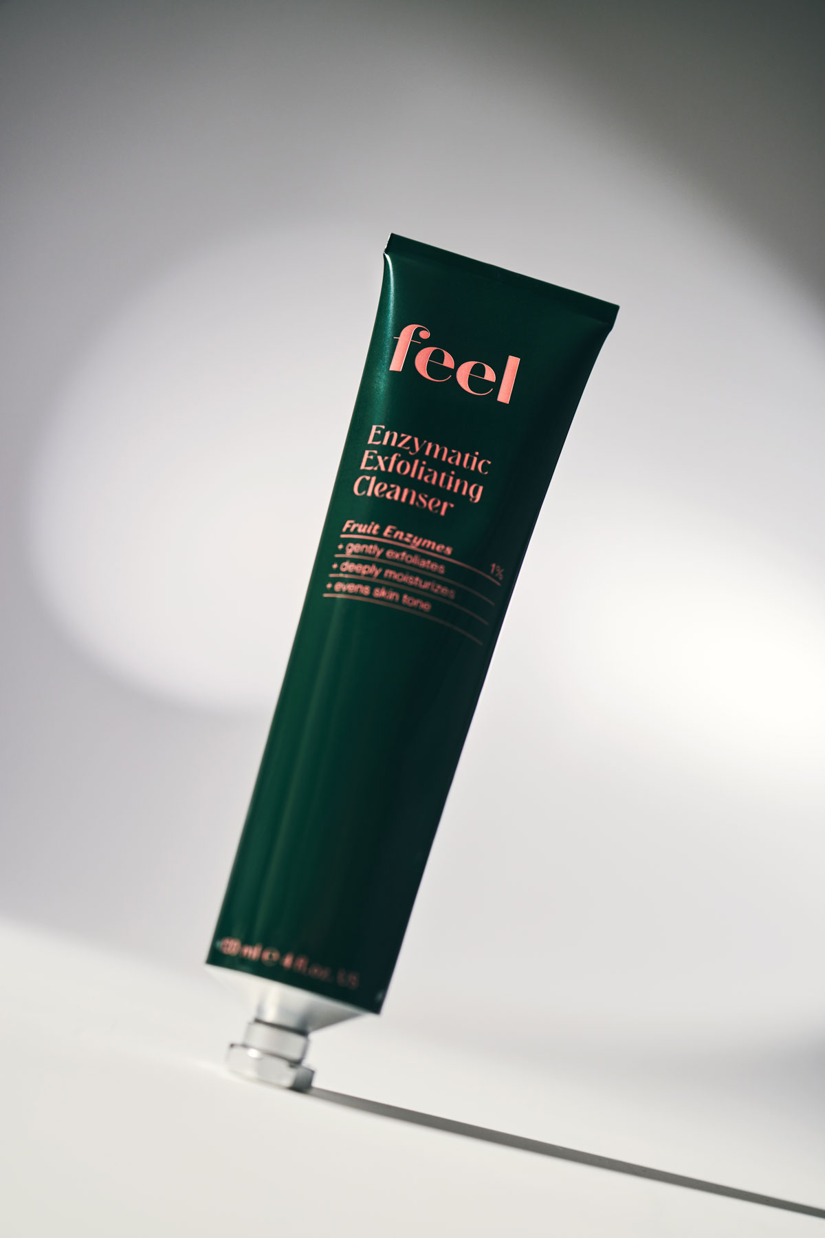 A dark green aluminum tube that says "Enzymatic Exfoliating Cleanser" stands in the centre of a grey background. The image is taken at an angle.