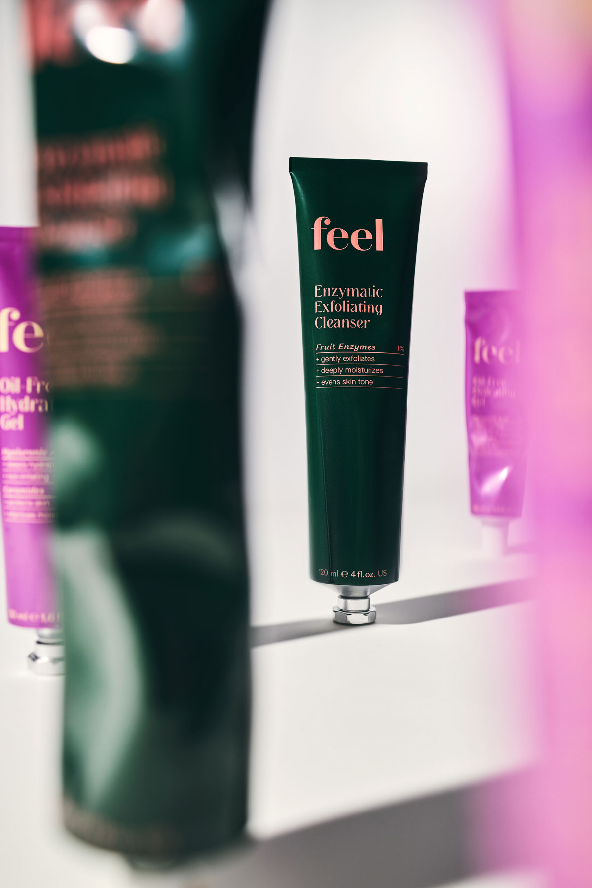A dark green aluminum tube stands in the centre in focus and says "Enzymatic Exfoliating Cleanser" surrounded by tubes standing in the foreground and distance out of focus and blurred